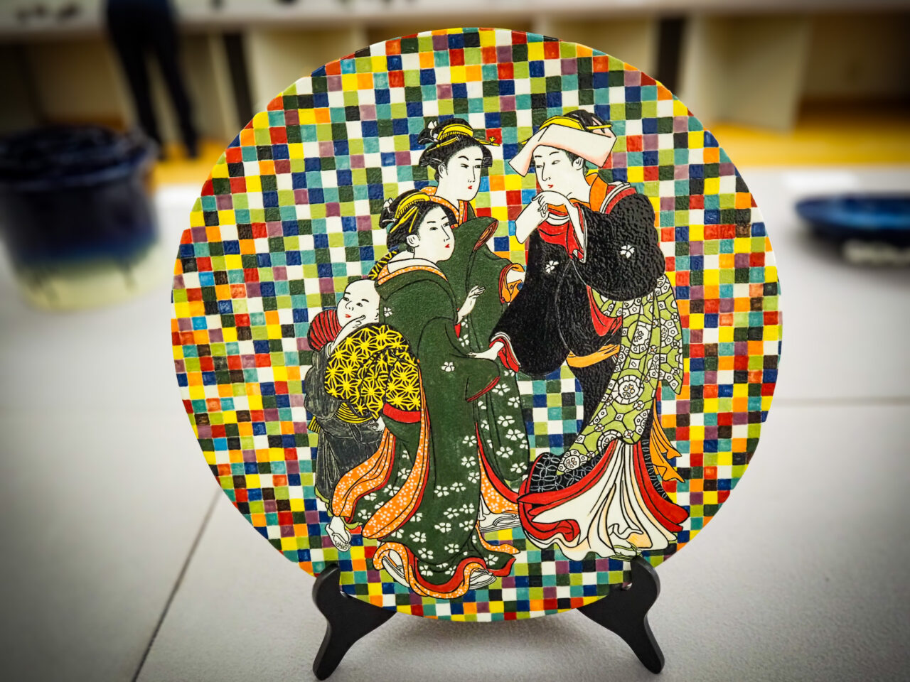 市松模様の背景に浮世絵風の女性たちが描かれた丸型の陶芸作品